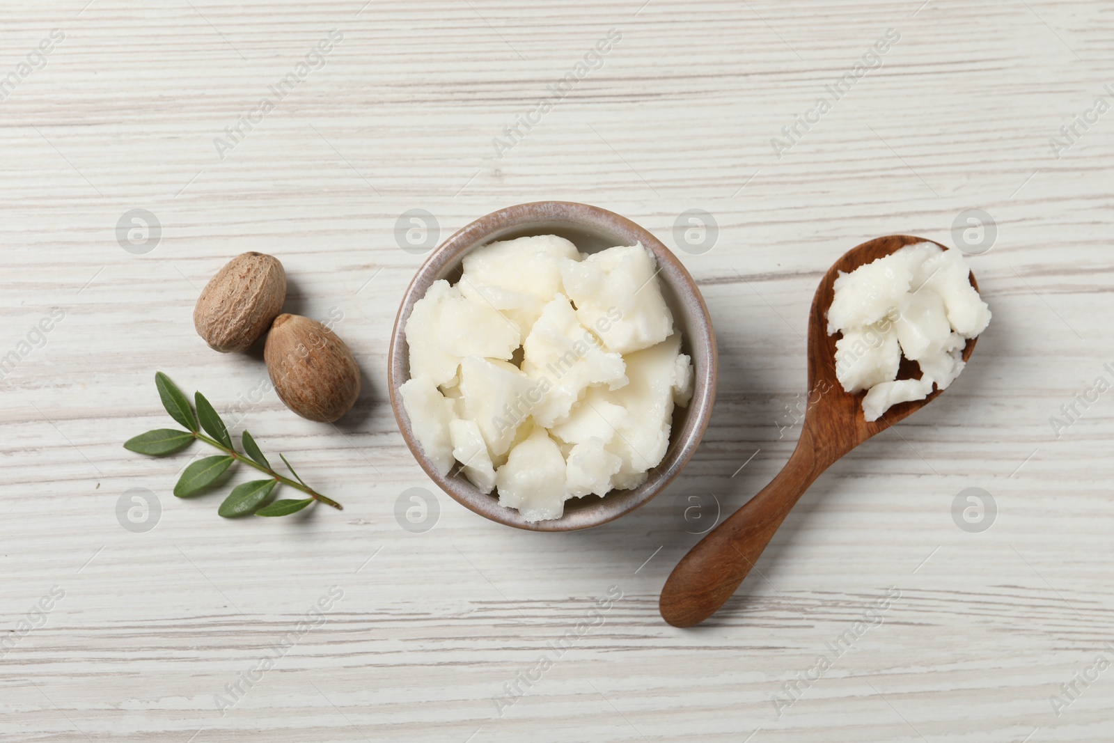 Photo of Shea butter in bowl, spoon and nuts on white wooden table, flat lay