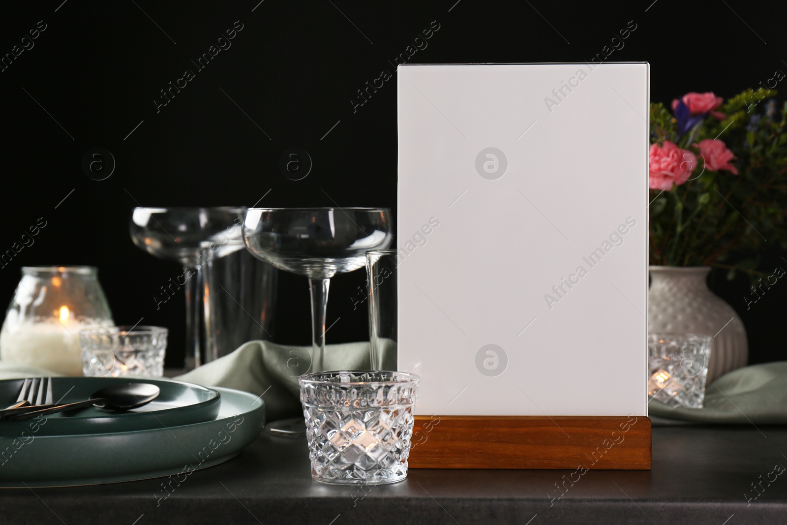 Photo of Menu holder, clean dishware and burning candles on grey table against black background