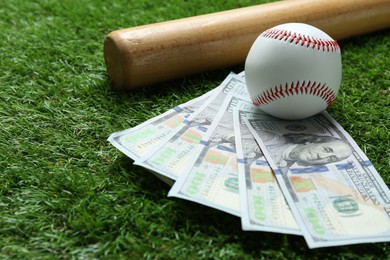 Photo of Baseball ball, bat and dollar banknotes on green grass outdoors, closeup