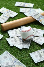 Photo of Baseball ball, bat and dollar banknotes on green grass outdoors