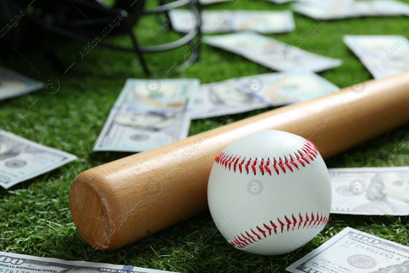 Photo of Baseball ball, bat and dollar banknotes on green grass outdoors, closeup