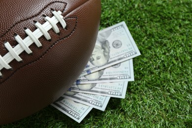 Photo of American football ball and dollar banknotes on green grass outdoors, closeup