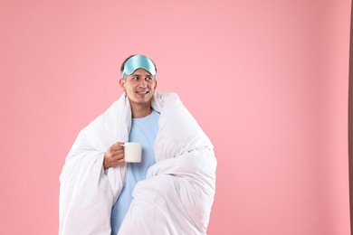 Photo of Happy man with sleep mask and cup wrapped in blanket on pink background, space for text