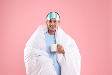 Photo of Happy man with sleep mask and cup wrapped in blanket on pink background