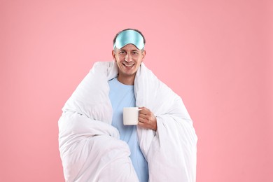 Happy man with sleep mask and cup wrapped in blanket on pink background