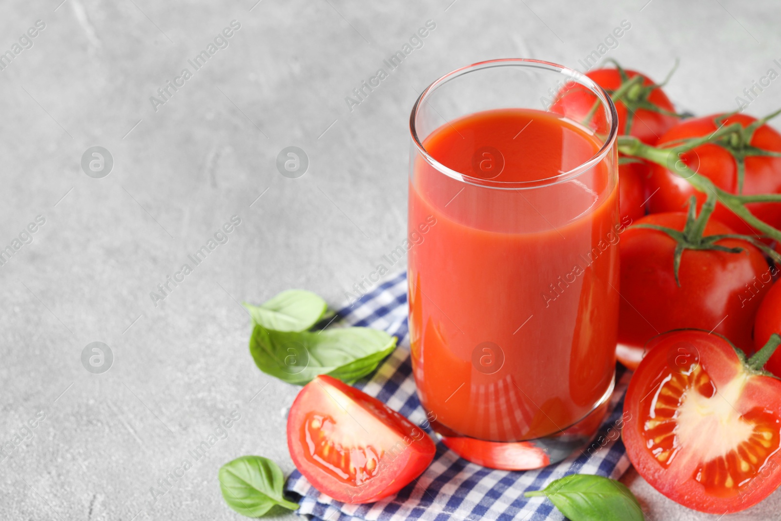 Photo of Tasty tomato juice in glass, basil leaves and fresh vegetables on grey textured table, space for text