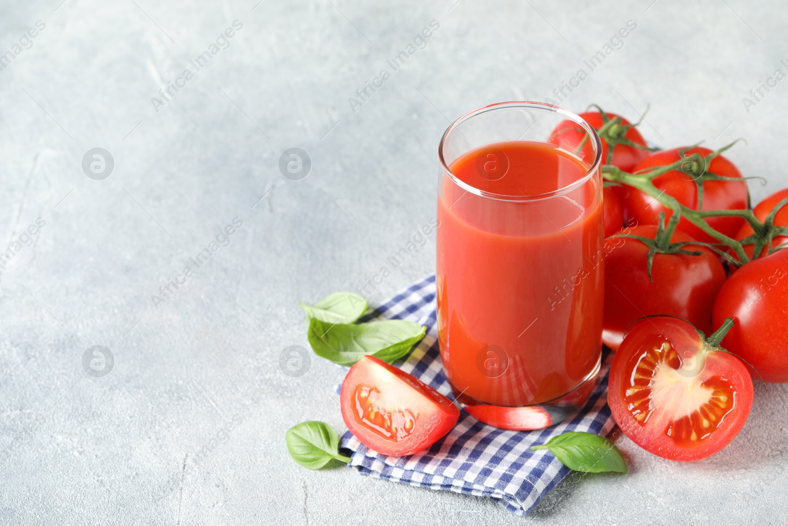 Photo of Tasty tomato juice in glass, basil leaves and fresh vegetables on grey textured table, space for text