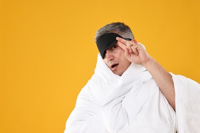 Photo of Overslept man with sleep mask wrapped in blanket on orange background, space for text