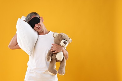 Photo of Overslept man with sleep mask, pillow and teddy bear on orange background, space for text