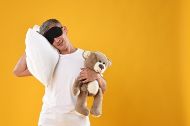 Overslept man with sleep mask, pillow and teddy bear on orange background, space for text