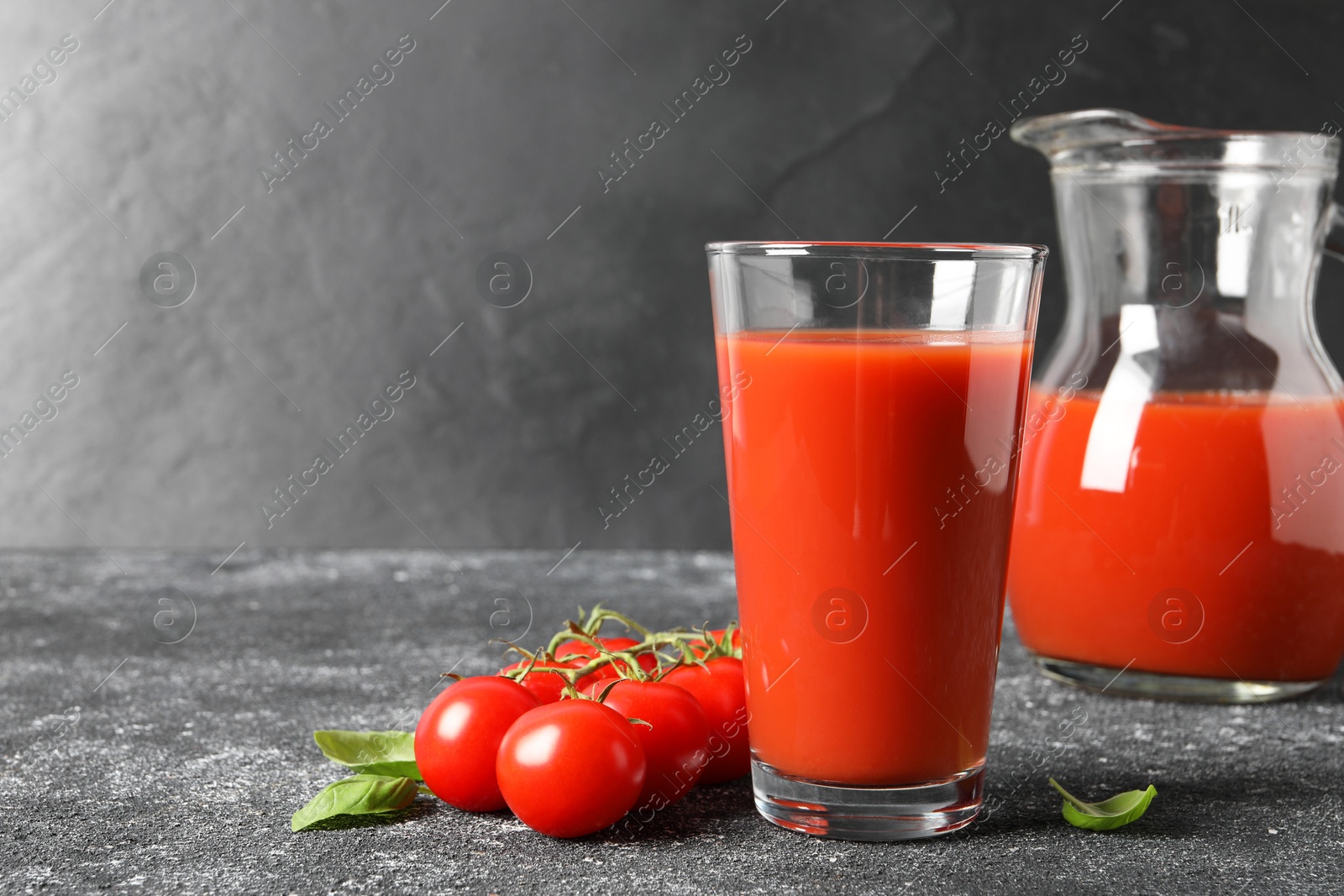 Photo of Tasty tomato juice in glass, jug, basil and fresh vegetables on grey table. Space for text