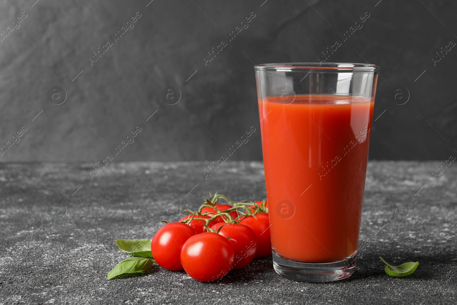 Photo of Tasty tomato juice in glass, basil and fresh vegetables on grey table. Space for text