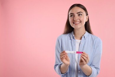 Photo of Happy woman holding pregnancy test on pink background, space for text