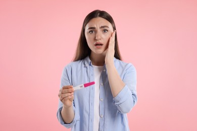 Shocked woman holding pregnancy test on pink background