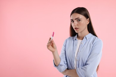 Confused woman holding pregnancy test on pink background, space for text