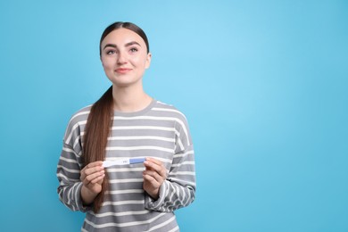 Happy woman holding pregnancy test on light blue background, space for text