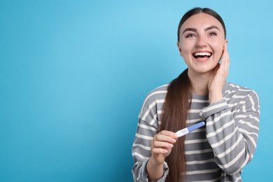 Happy woman holding pregnancy test on light blue background, space for text