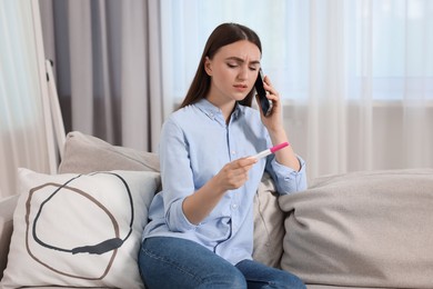 Sad woman with pregnancy test talking on smartphone indoors