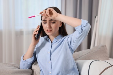 Sad woman with pregnancy test talking on smartphone indoors