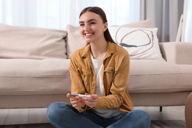 Happy woman holding pregnancy test on floor indoors