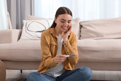 Happy woman holding pregnancy test on floor indoors