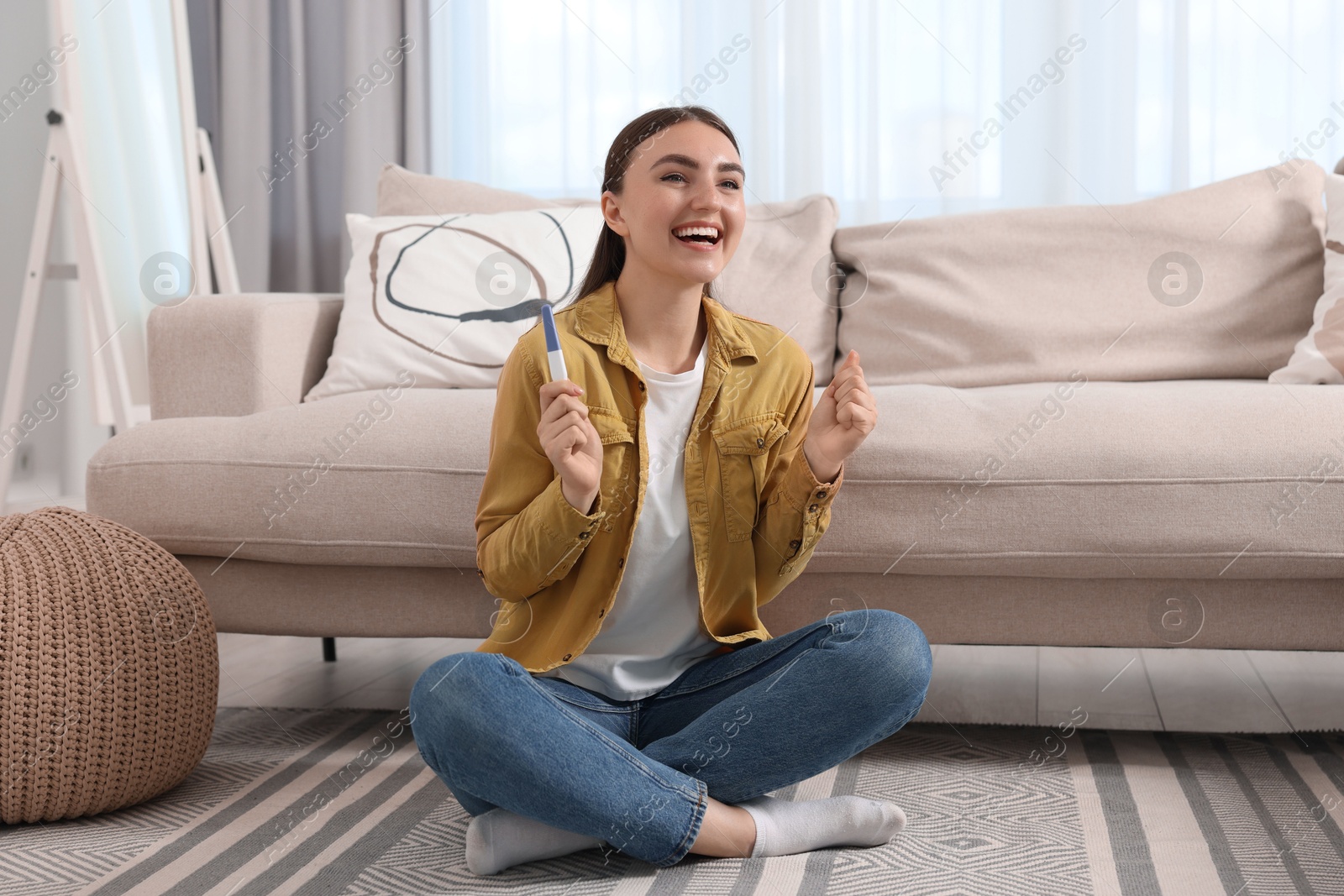 Photo of Happy woman holding pregnancy test on floor indoors