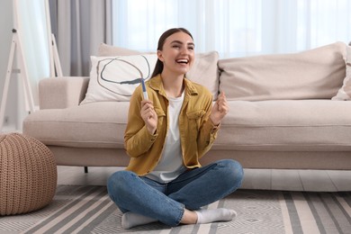 Happy woman holding pregnancy test on floor indoors