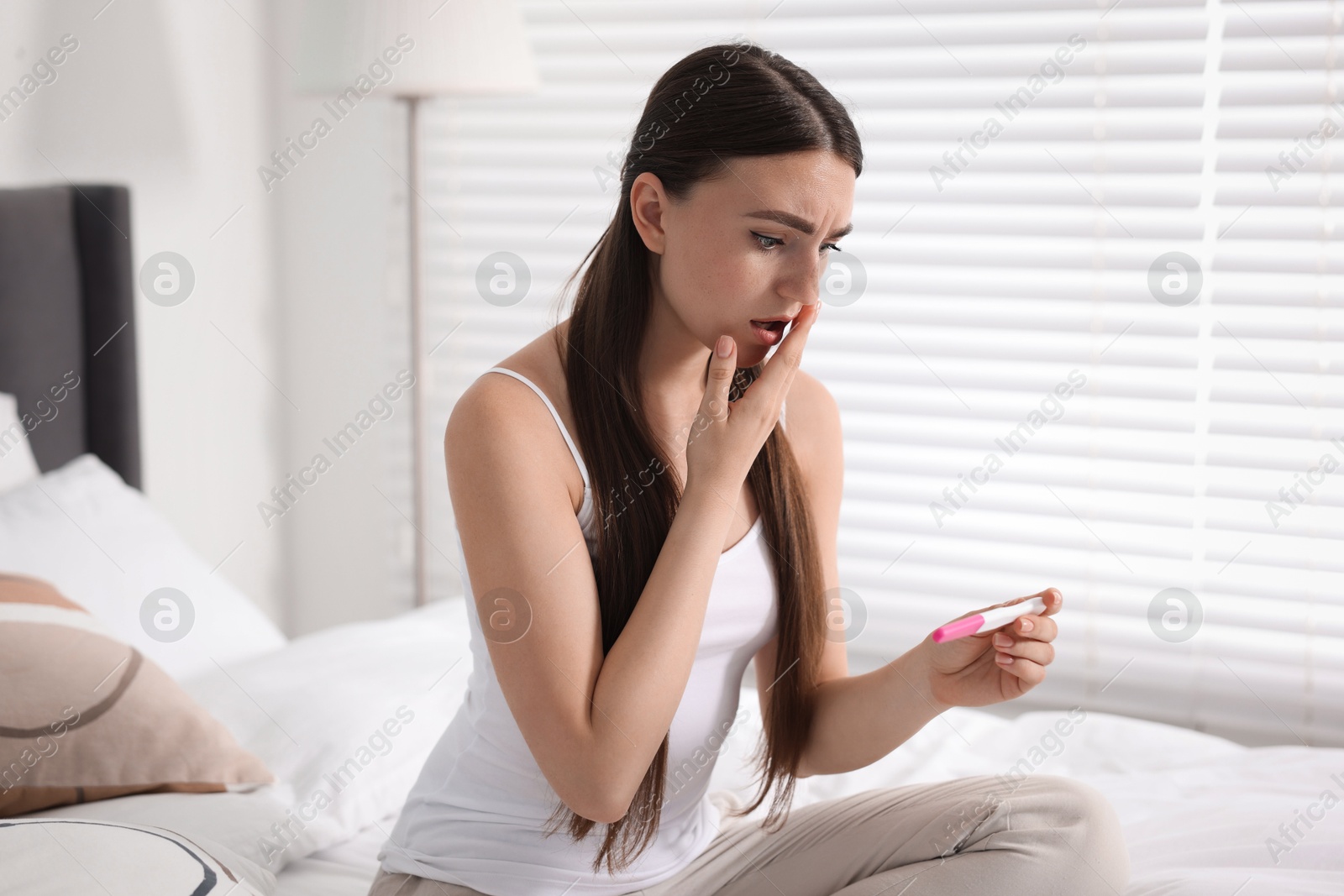 Photo of Shocked woman holding pregnancy test on bed in room