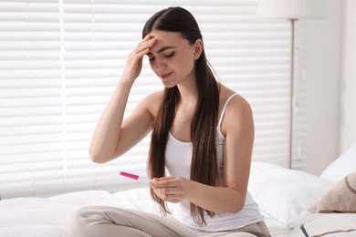 Sad woman holding pregnancy test on bed in room