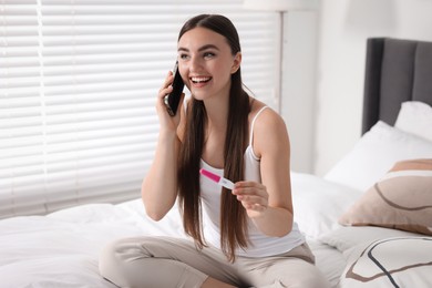 Photo of Happy woman with pregnancy test talking on smartphone in room