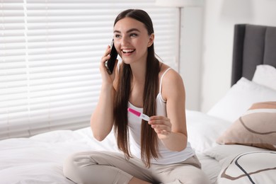 Happy woman with pregnancy test talking on smartphone in room