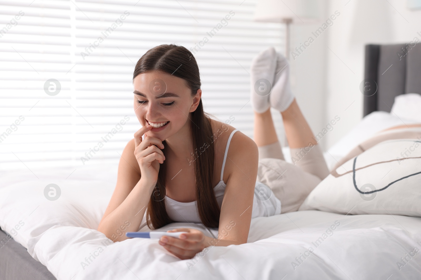 Photo of Happy woman holding pregnancy test on bed in room