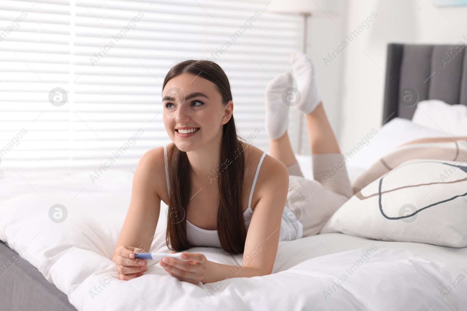 Photo of Happy woman holding pregnancy test on bed in room