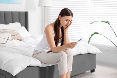 Sad woman holding pregnancy test on bed in room