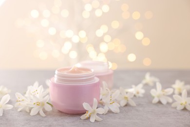 Photo of Cream in jars and beautiful jasmine flowers on grey table against beige background with blurred lights, closeup