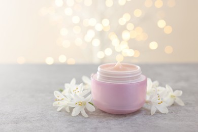 Photo of Cream in jar and beautiful jasmine flowers on grey table against light background with blurred lights, closeup