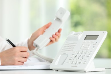 Assistant with telephone handset writing at white table against blurred background, closeup