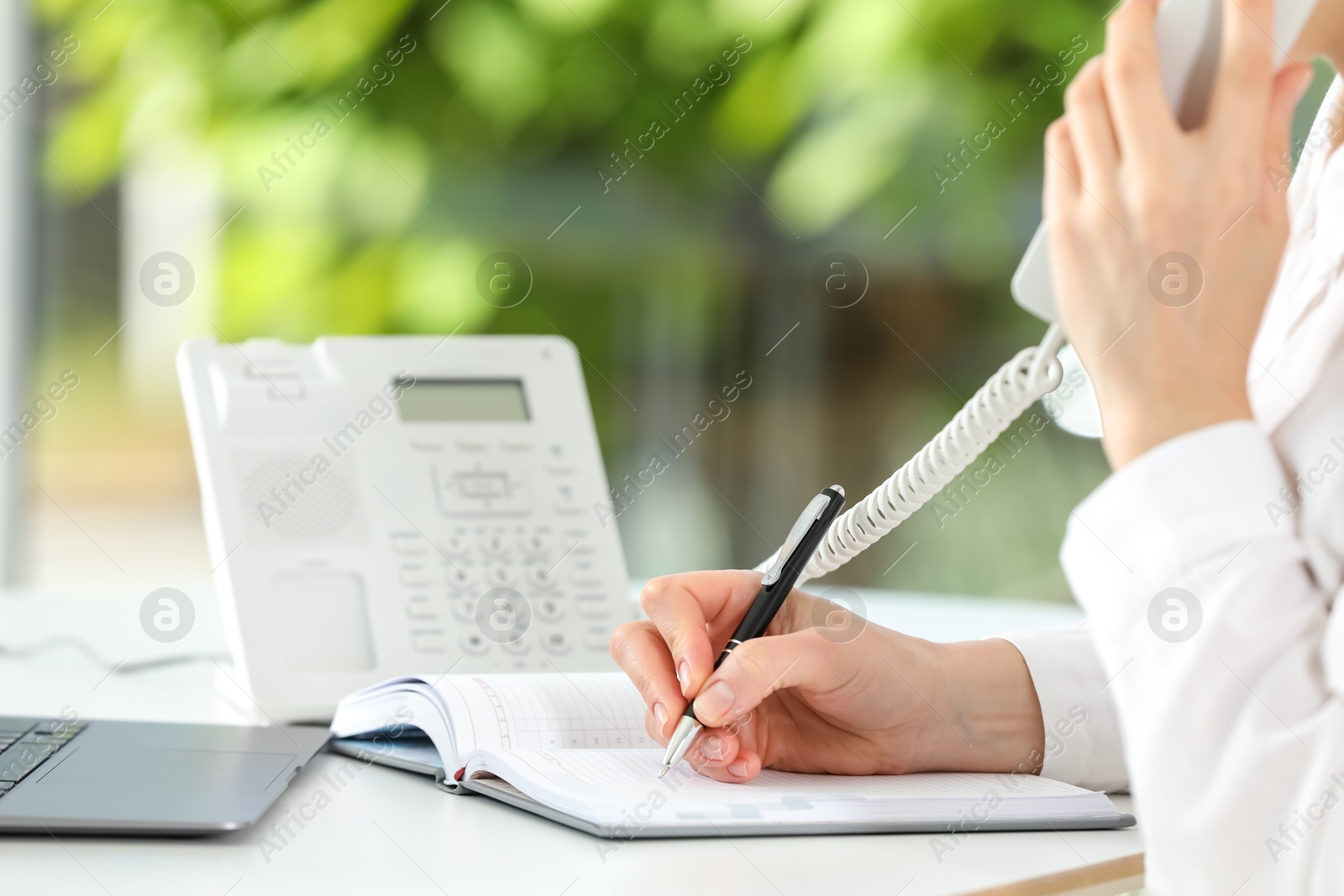 Photo of Assistant with telephone handset writing at white table against blurred green background, closeup