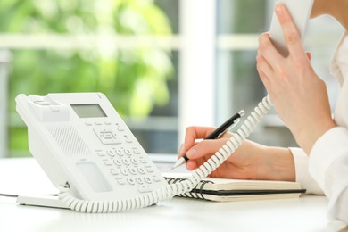 Assistant with telephone handset writing at white table against blurred green background, closeup