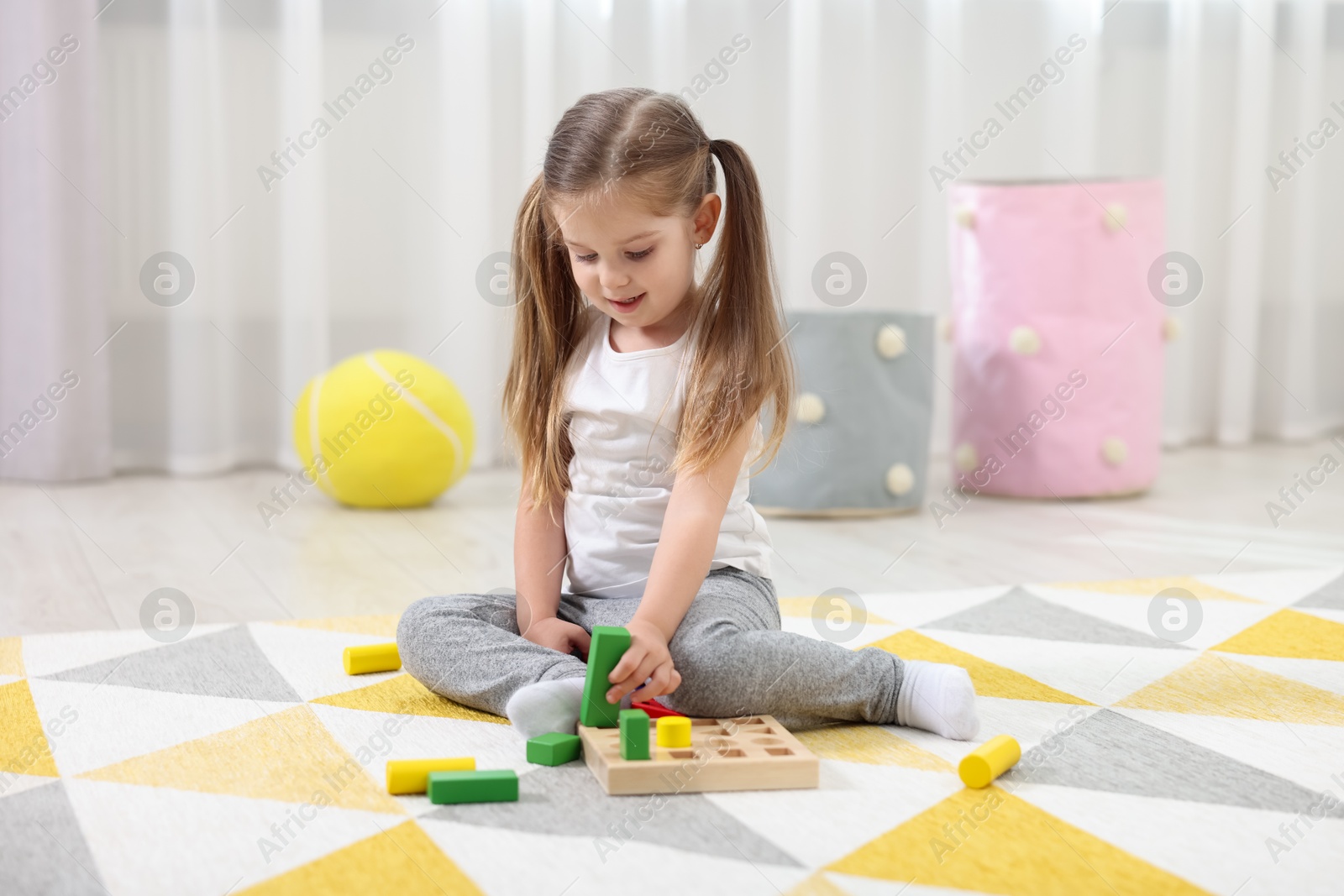 Photo of Cute little girl playing with set of wooden geometric figures on floor in kindergarten