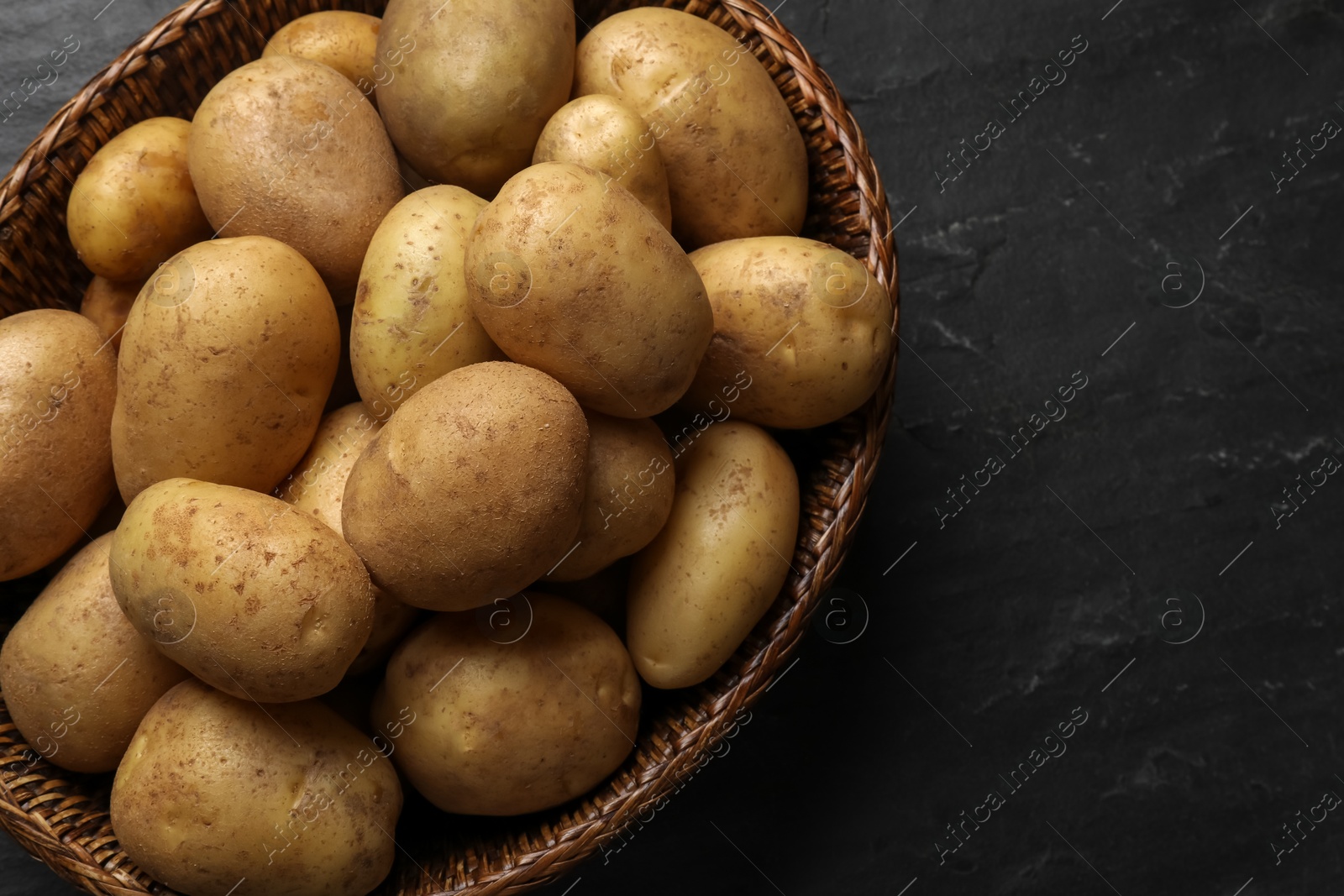 Photo of Many fresh potatoes in wicker basket on black textured table, top view. Space for text