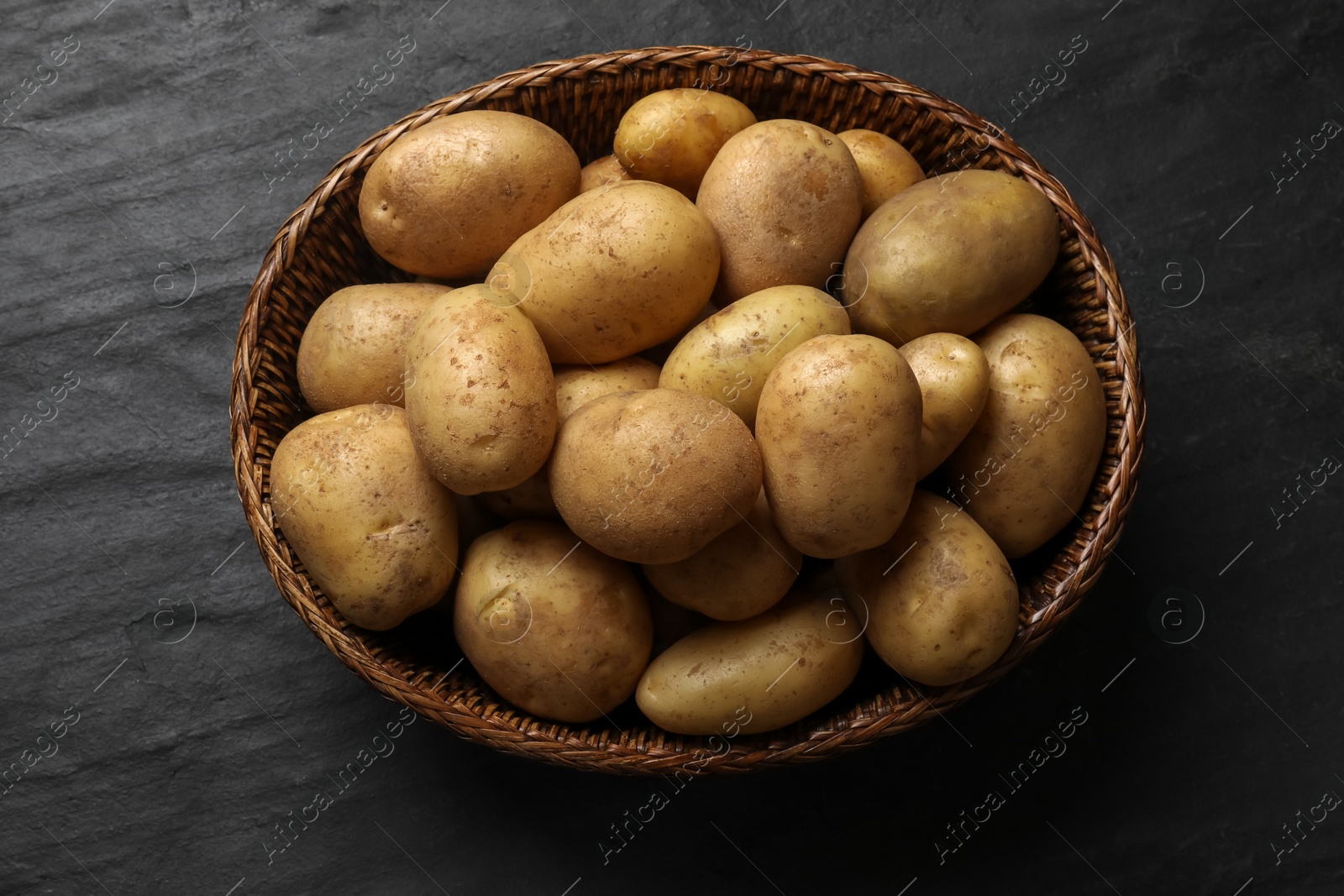 Photo of Many fresh potatoes in wicker basket on black textured table, top view