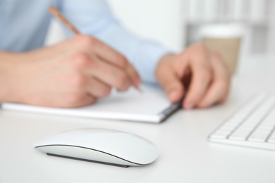 Photo of Wireless mouse on white table, closeup. Man writing in notebook indoors, selective focus