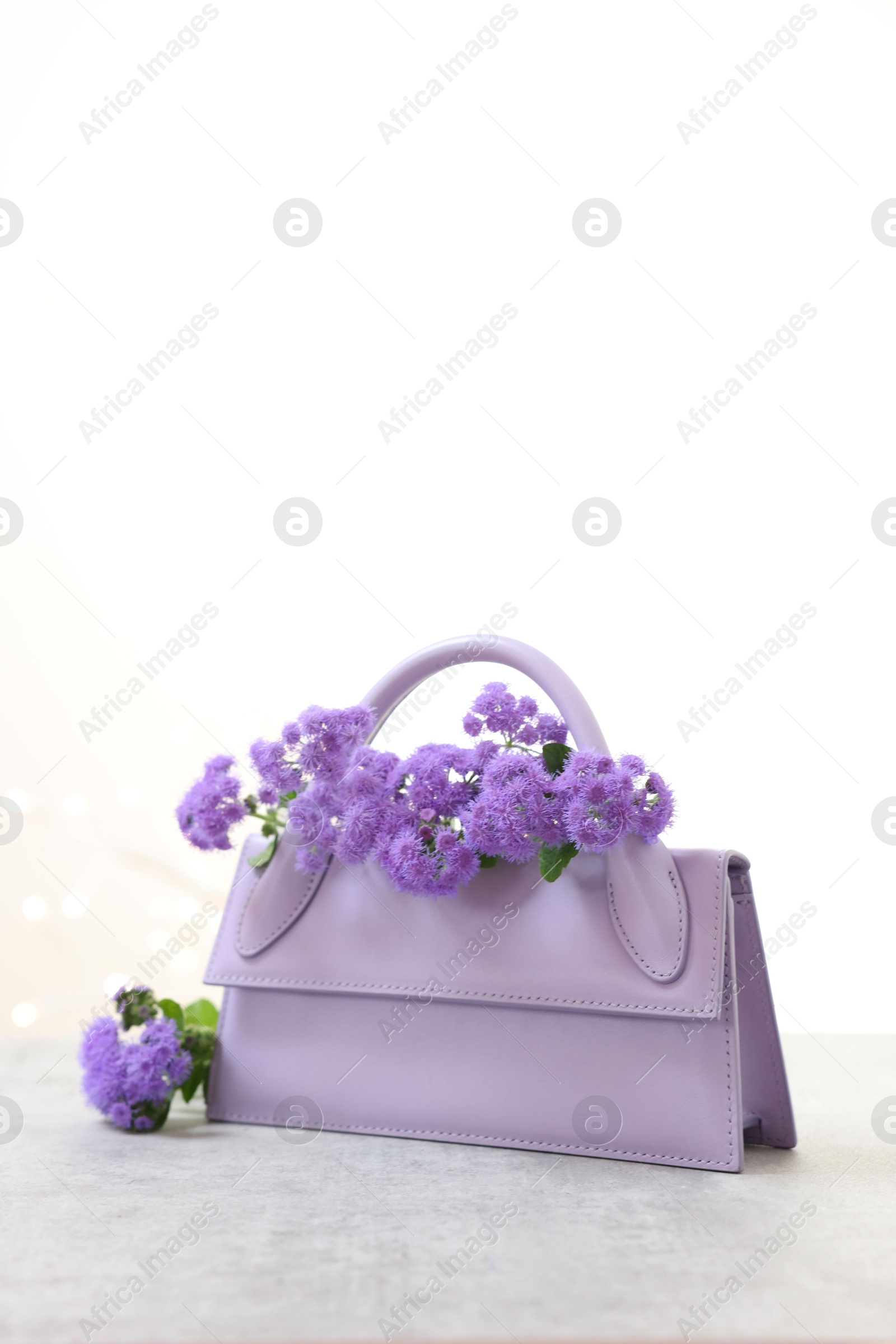 Photo of Stylish violet bag and ageratum flowers on gray table against light background, space for text