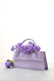 Stylish violet bag and ageratum flowers on gray table against light background, space for text