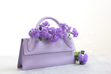 Photo of Stylish violet bag and ageratum flowers on gray table against light background