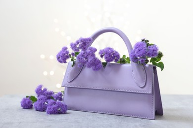 Photo of Stylish violet bag and ageratum flowers on gray table against light background