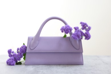 Photo of Stylish violet bag and ageratum flowers on gray table against light background