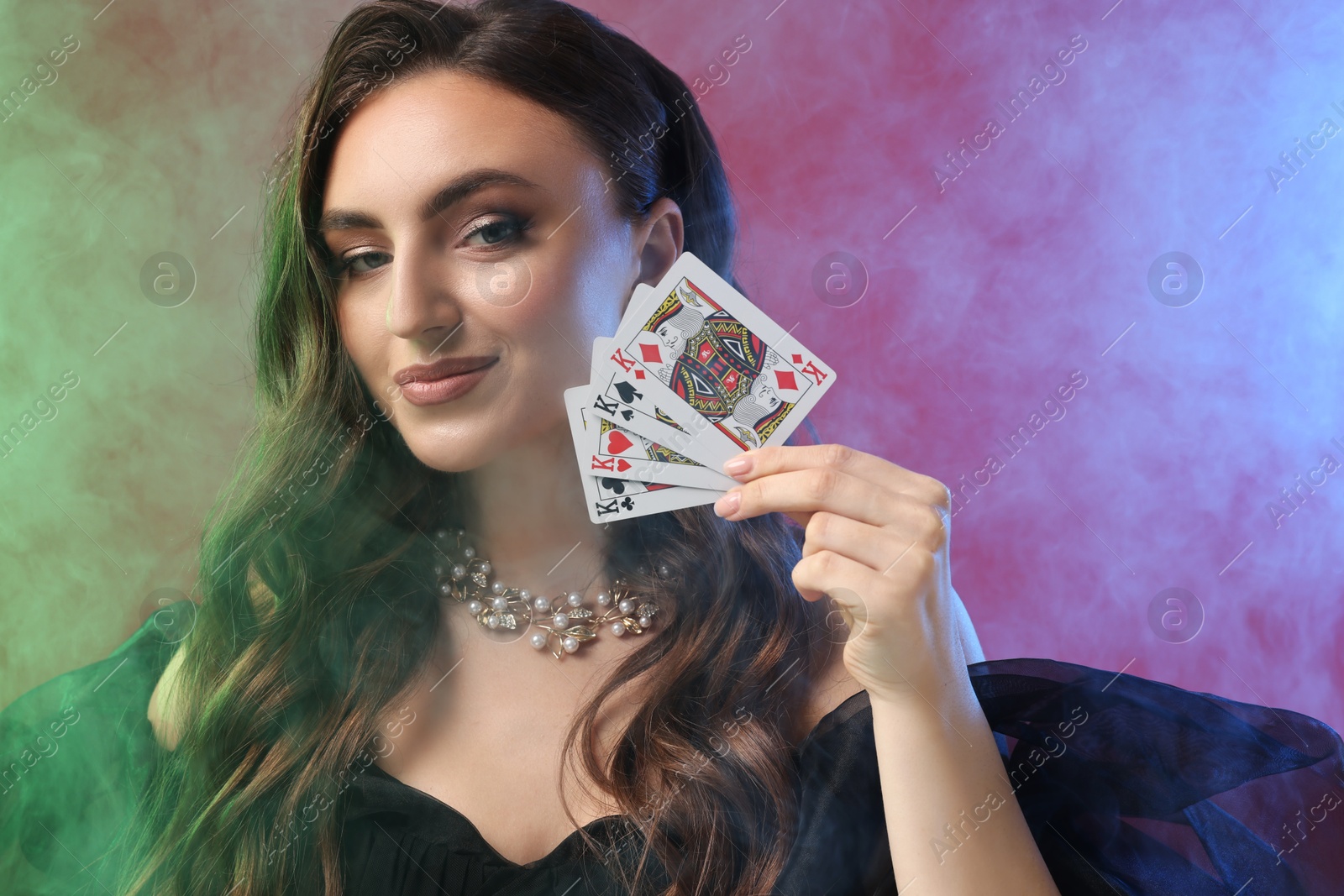 Photo of Poker game. Charming woman with playing cards on red background in color lights and smoke