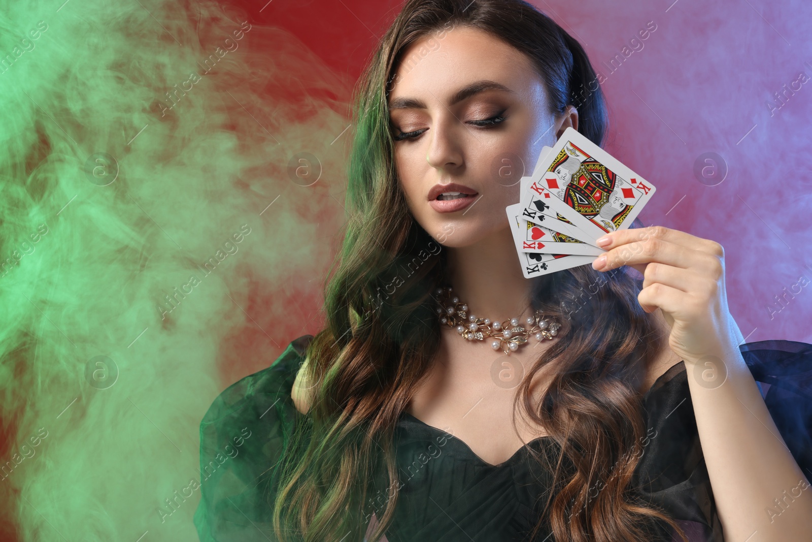 Photo of Poker game. Charming woman with playing cards on red background in color lights and smoke